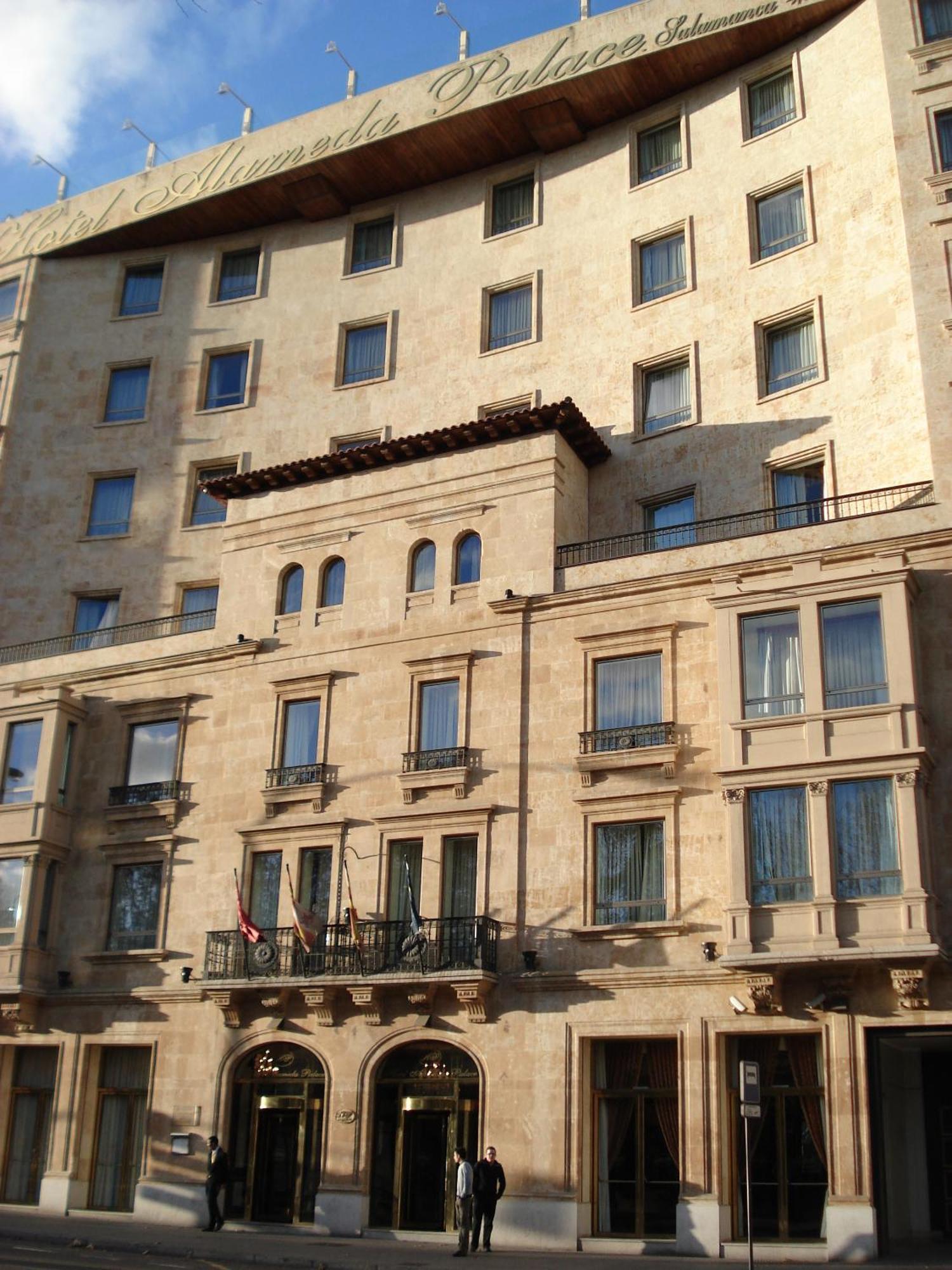 Hotel Alameda Palace Salamanca Exterior foto The photo depicts a building with a stone facade that features a mix of architectural designs. The lower section includes several large windows framed by decorative elements, with balconies that have railings. There are also flags visible at the entr